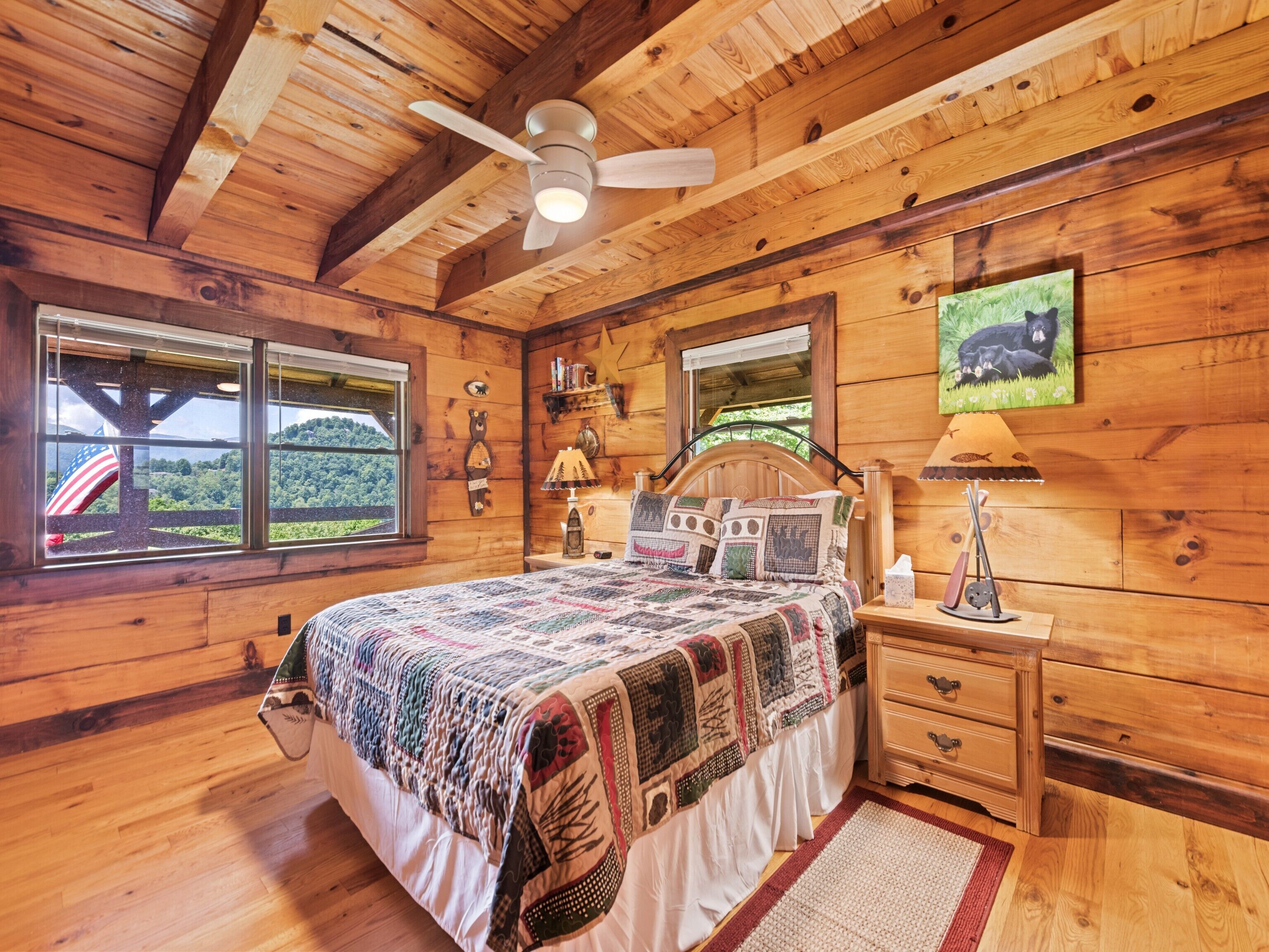 Ein Schlafzimmer in einer Holzhütte mit einem Bett, einem Nachttisch, einer Lampe, einem Deckenventilator und einem Fenster mit Blick ins Freie.