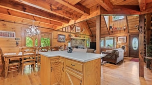 A rustic cabin interior with a wooden dining area, a kitchen island, a living room with a sofa, and a stone fireplace. - 4 Bd 3 Ba Log Cabin, Private, Views, Close to Town, Game, Hot Tub, Firepit (Waynesville)