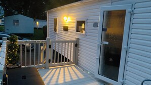 Una casa móvil blanca con un porche, una puerta corredera de vidrio y una pequeña terraza decorada con luces colgantes.