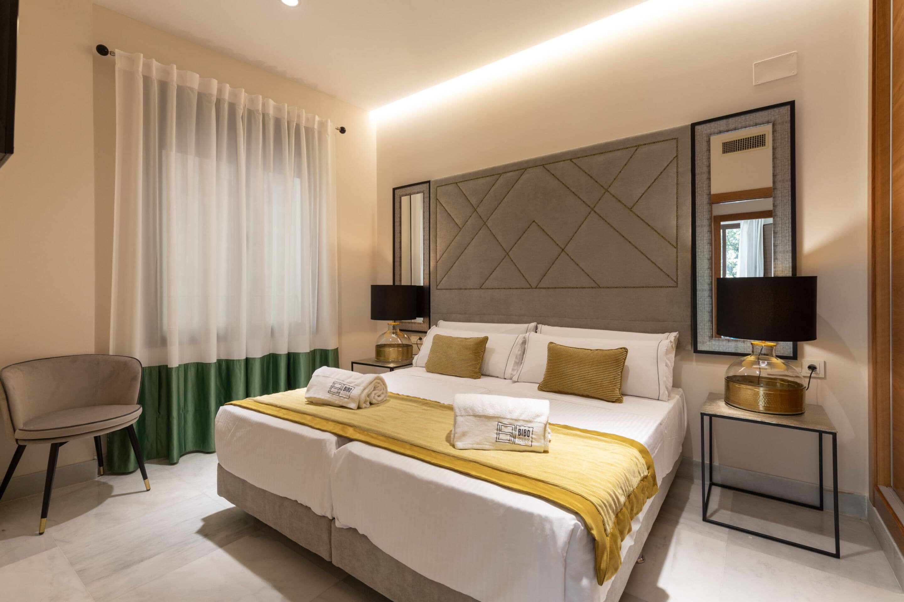 Una habitación de hotel con una cama doble, dos mesitas de noche con lámparas, una silla y un ventanal con cortinas translúcidas.