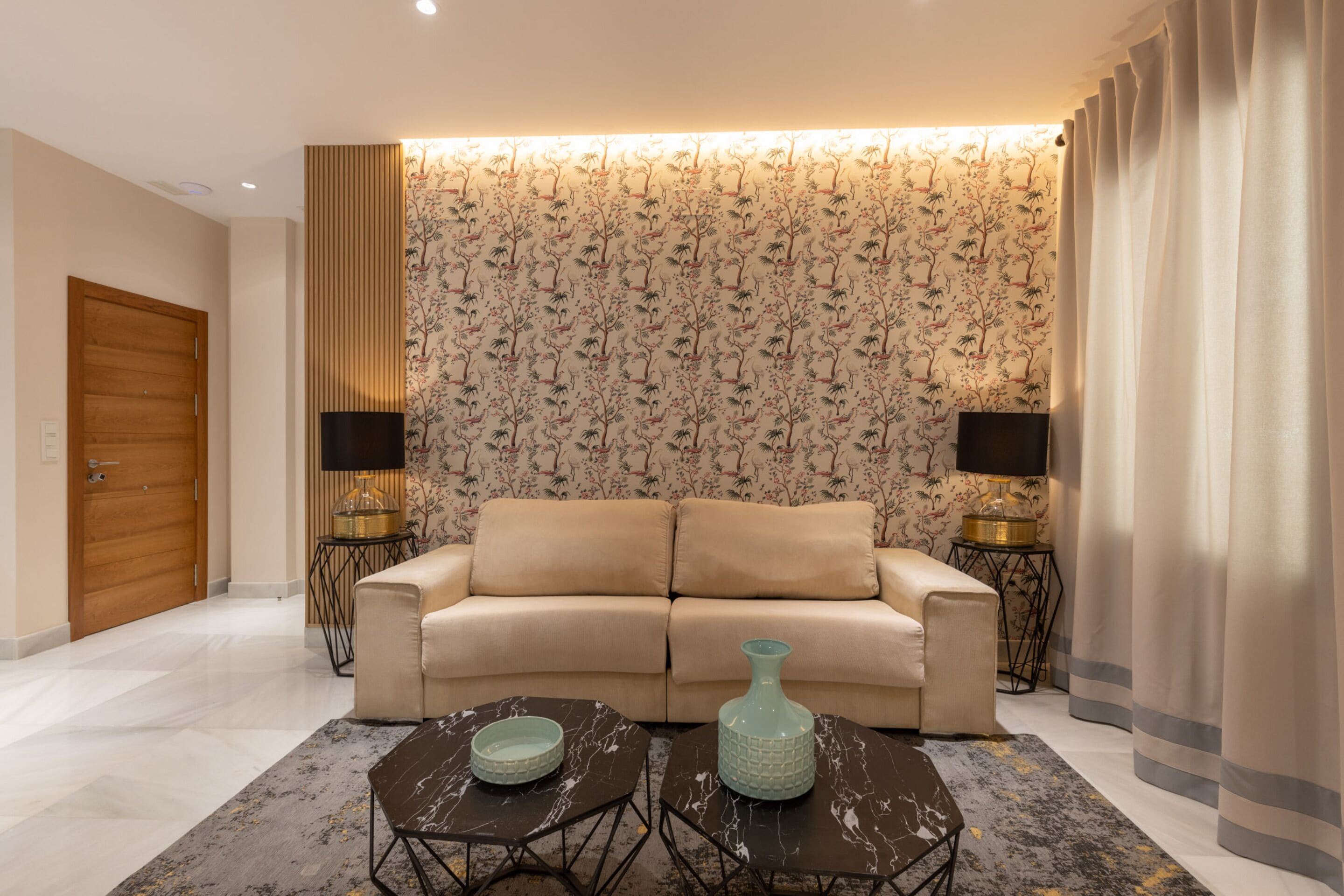 A living room with a beige sofa, two side tables with lamps, a patterned wallpaper, and a large window with curtains.