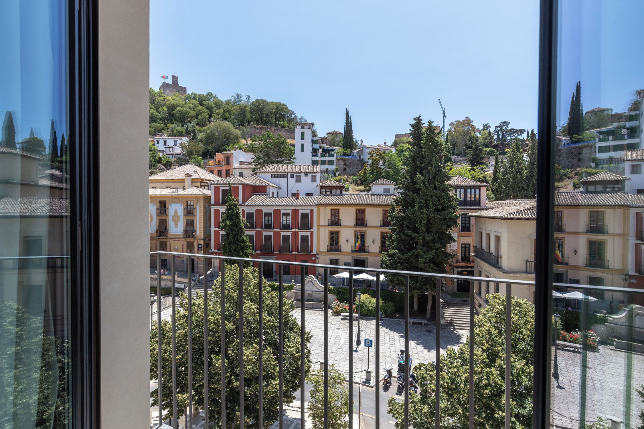 Vista des d'una finestra sobre una plaça amb edificis, arbres i un cel serè.