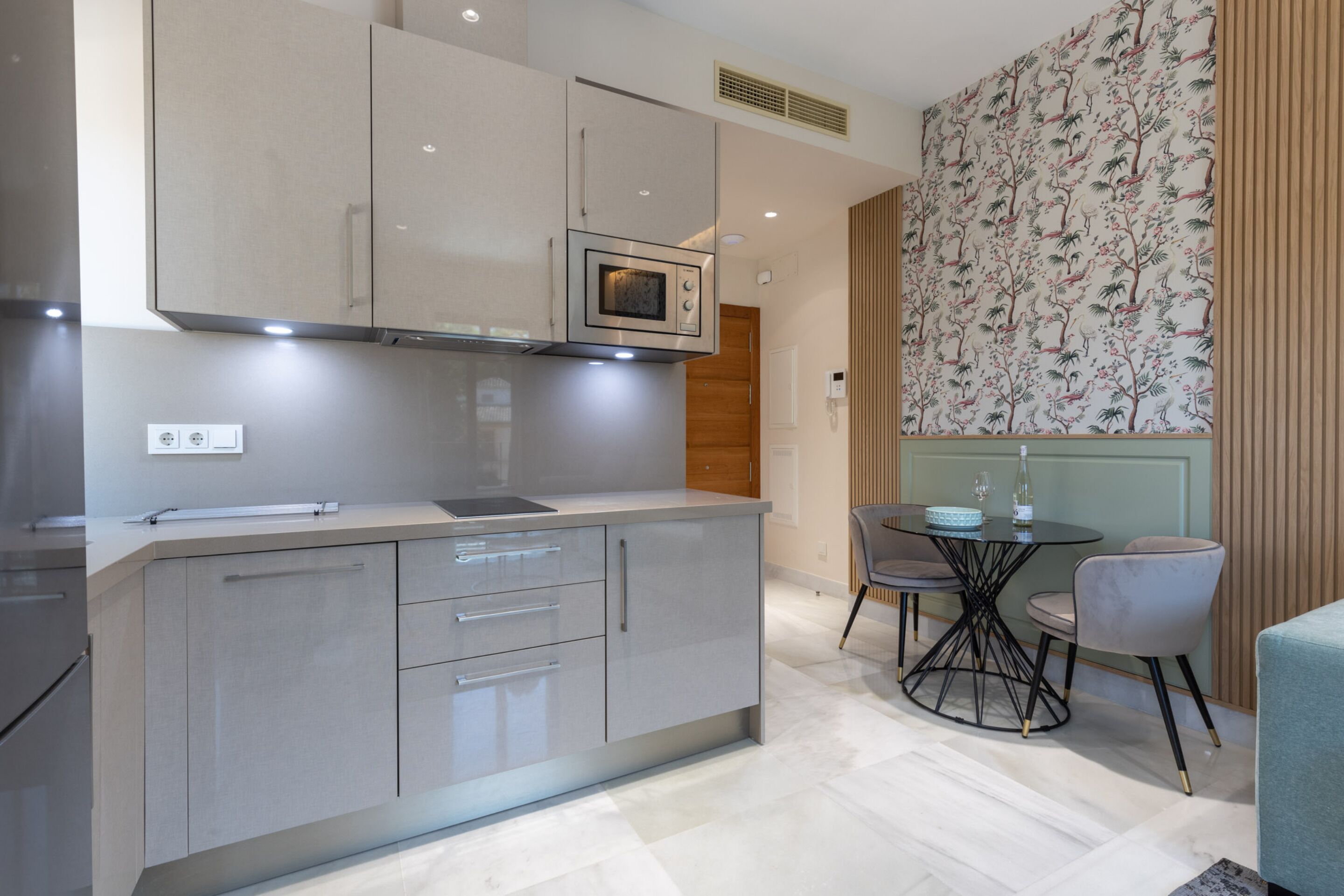 A modern kitchen with a built-in microwave, a dining area with a round table and chairs, and a decorative wall with a floral pattern.