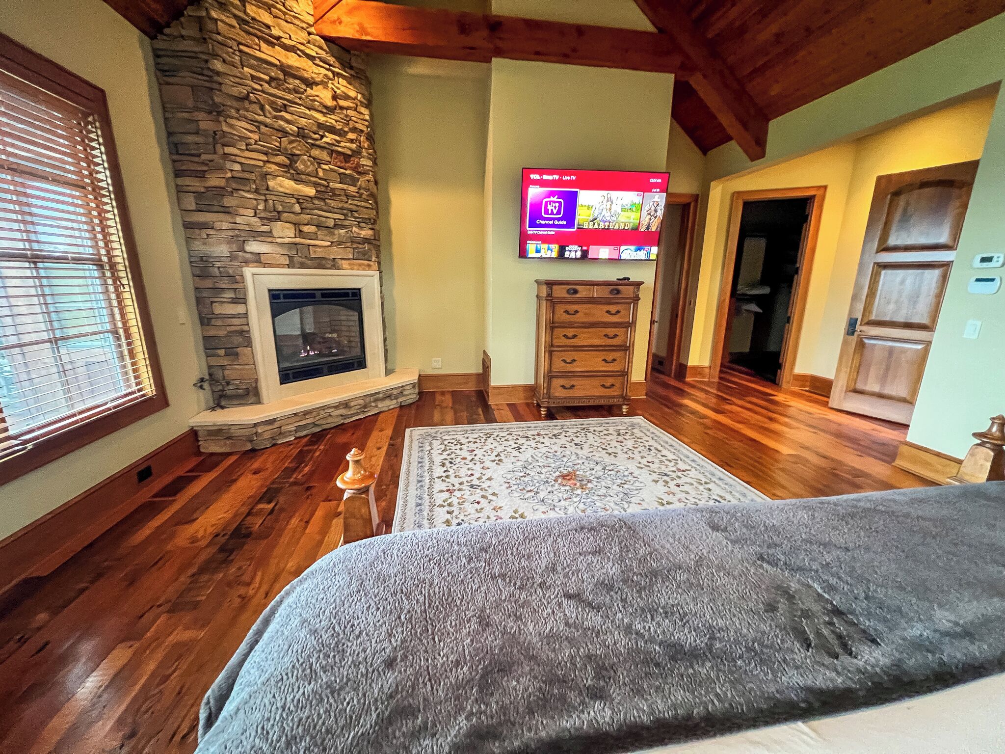 A cozy living room with a stone fireplace, wooden floors, a television mounted on the wall, and a large rug.