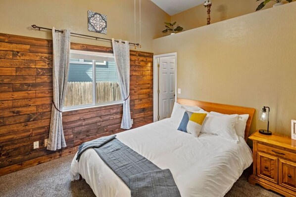 A bedroom with a wooden wall, a bed with white bedding, a nightstand with a lamp, and a window with curtains. - Family Mountain Retreat ~Game Table ~Fire Table (Flagstaff)