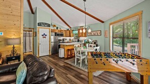 A cozy living room with a foosball table, a dining area, and a kitchen with wooden cabinets and a hanging light fixture.