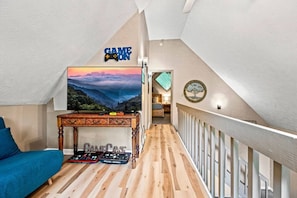 A cozy loft bedroom with a sloped ceiling, a large flat-screen TV, a wooden desk with DJ equipment, and a blue sofa.