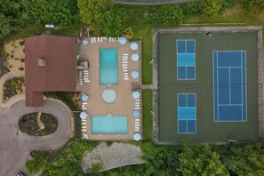 Eine Vogelperspektive zeigt ein Anwesen mit Schwimmbad, Liegestühlen, einem Tennisplatz und einem Gebäude mit rotem Dach.