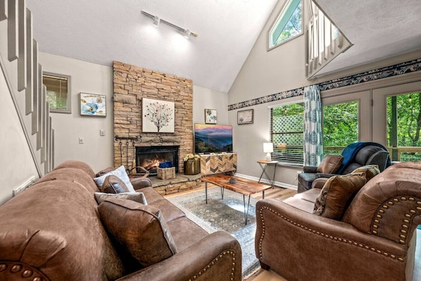 A spacious living room with a stone fireplace, leather sofas, and a large window offering a view of greenery.