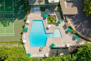 An aerial view of a swimming pool area with multiple sun loungers and umbrellas, surrounded by greenery and adjacent to tennis courts.