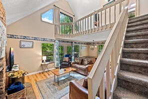A spacious living area with a high ceiling, a staircase, and a view of greenery outside.