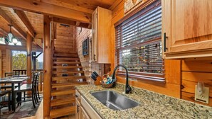 Interior de una cabaña de madera con área de cocina que incluye una encimera de granito, pileta de acero inoxidable y muebles de madera.