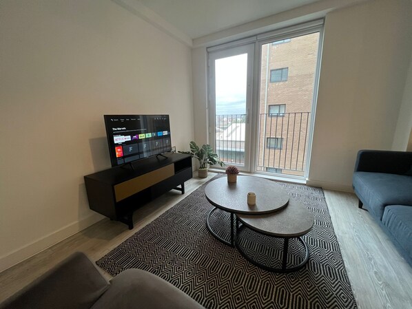 Un soggiorno moderno con una televisione a schermo piatto che mostra l'interfaccia di un servizio di streaming, un divano, un tavolino da caffè e un'ampia finestra con vista su un edificio.