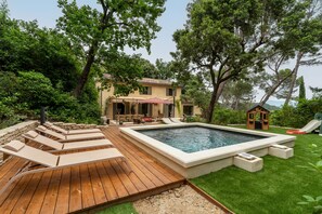 Un patio trasero con piscina, sillas de descanso, una terraza de madera y una casita de juegos.