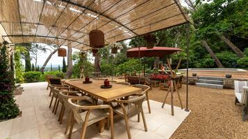 Une terrasse extérieure avec une table en bois et des chaises en osier, entourée d’une pergola ornée de jardinières suspendues.