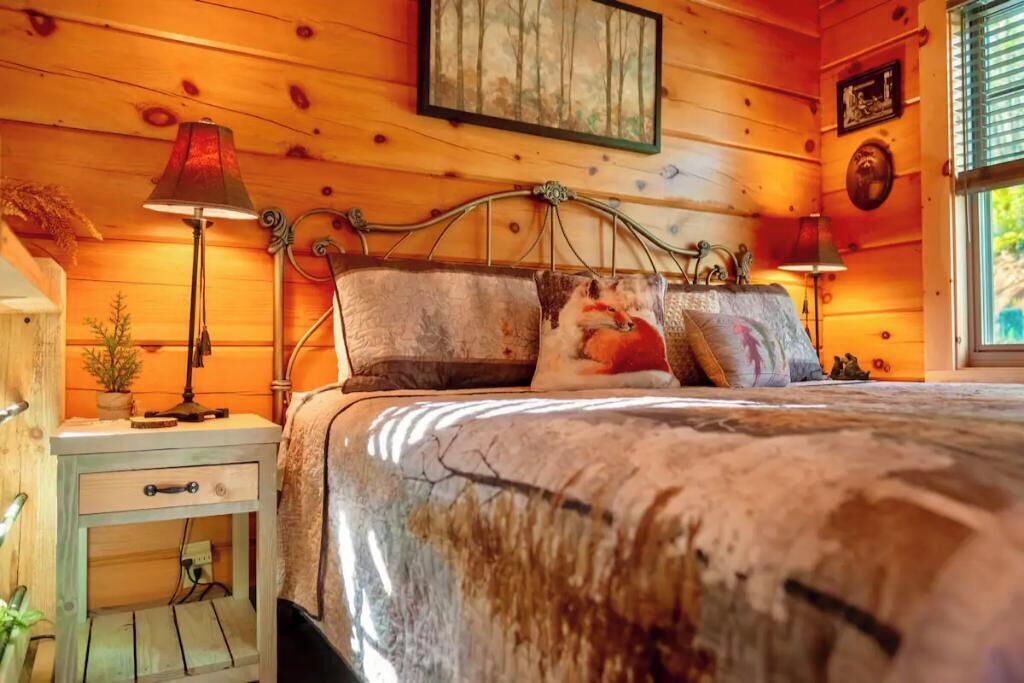 A wooden cabin bedroom with a metal bed frame, a nightstand with a lamp, and a window with blinds.