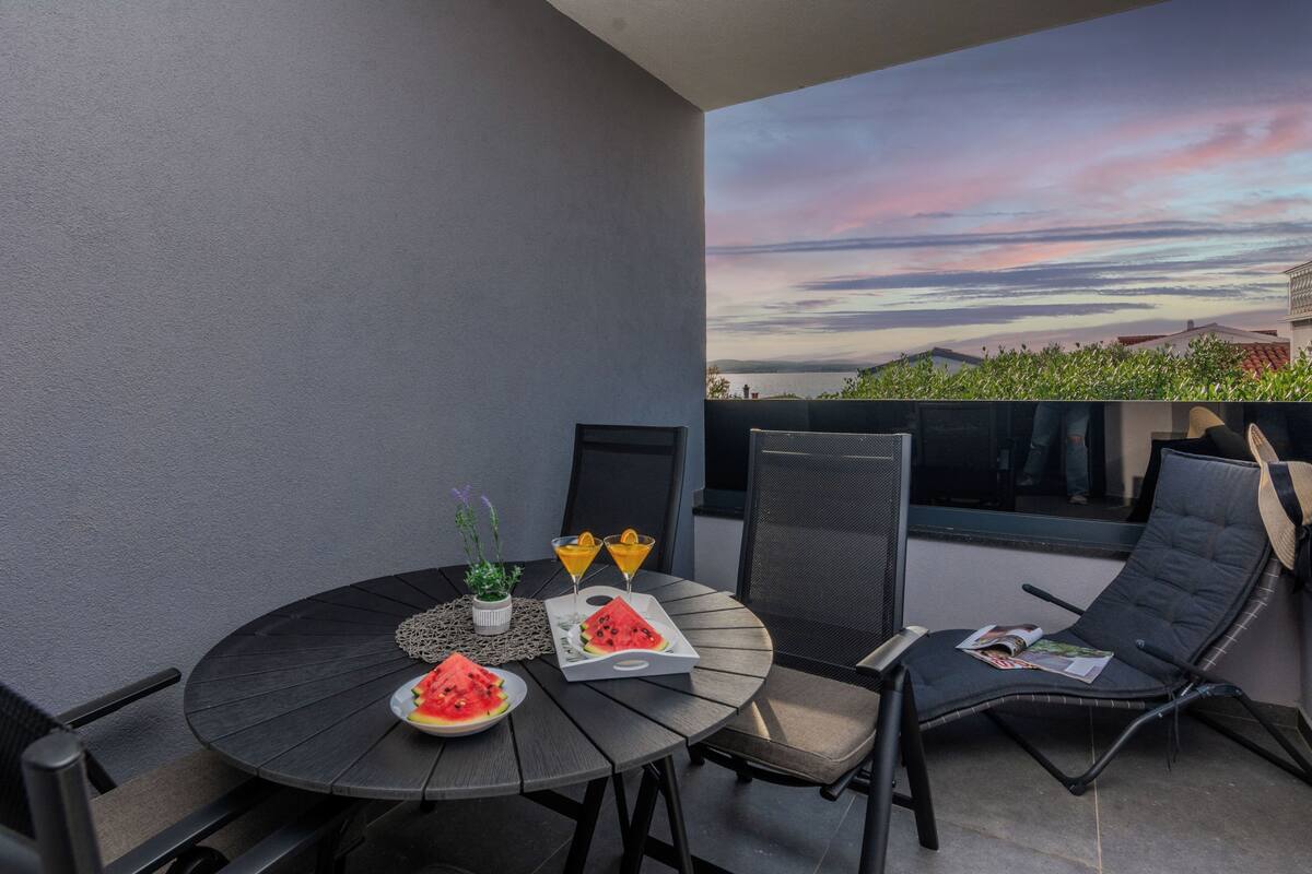 A round table with a tray of watermelon slices, two drinks, and a small plant. There are two black chairs and a lounge chair with a magazine.