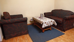 A room with two patterned sofas, a small wooden table with a floral tablecloth, and a blue rug on a wooden floor. - SSG COTTAGE (Ootacamund)