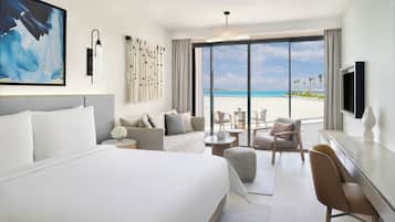 A modern hotel room with a large bed, a sofa, a desk, and a TV. The room has a large window with a view of the beach.