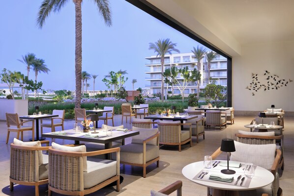 Un área de comedor al aire libre moderna con mobiliario de mimbre, mesas dispuestas con platos, vasos y cubiertos, con vistas a una piscina y palmeras.