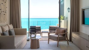 A modern hotel room with a large bed, a sofa, two armchairs, a small table, and a TV. The room has a large sliding glass door leading to a balcony with a view of the sea. - Address  Beach Resort Marassi (El Alamein)