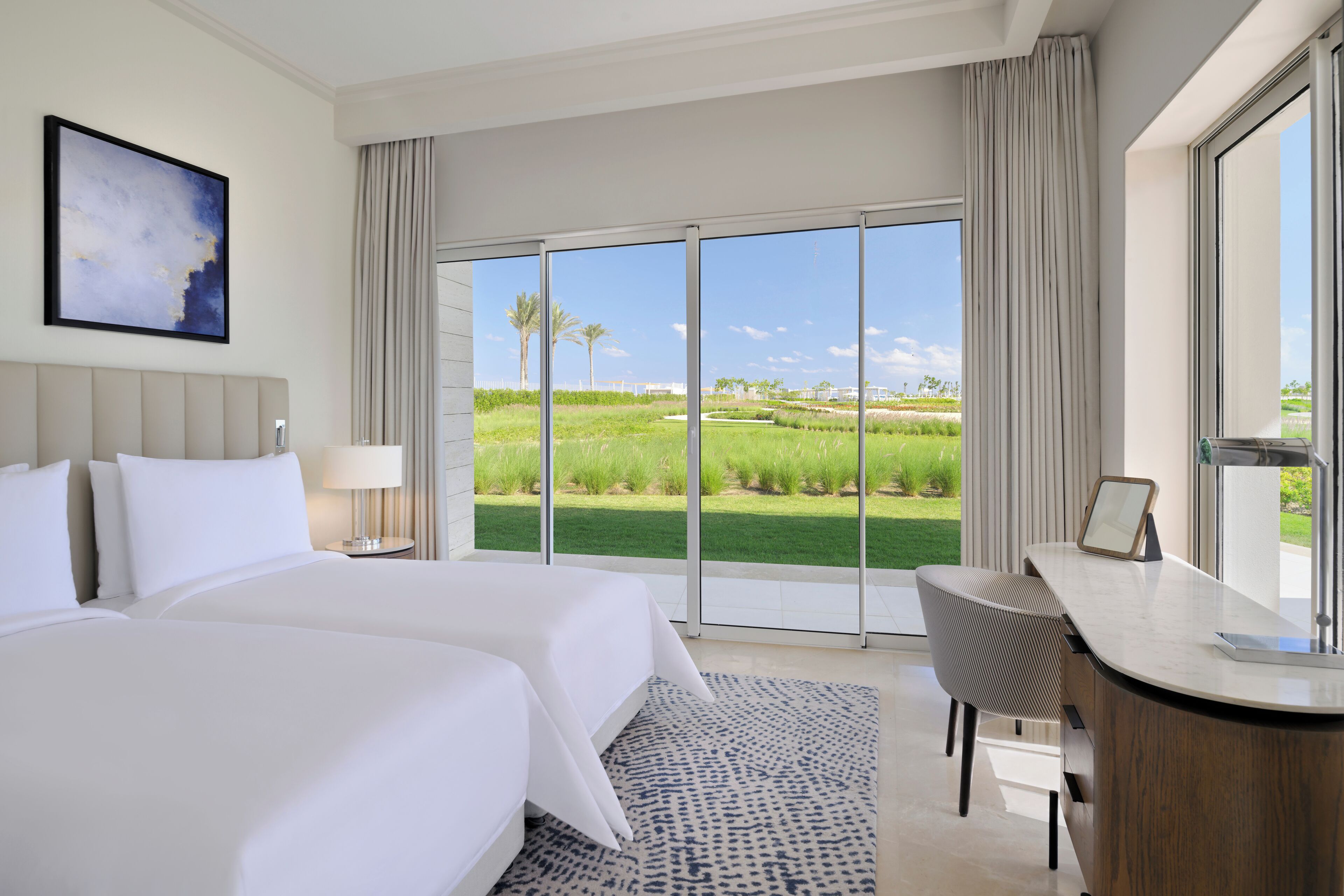A hotel room with a large bed, a desk, and a view of a green landscape through large windows.
