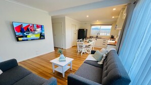 A modern living room with a TV, grey sofas, a small white table, and a dining area with a kitchen in the background. - Casa Del Mar: near beach/wineries/golf/Hot Springs (Tootgarook)
