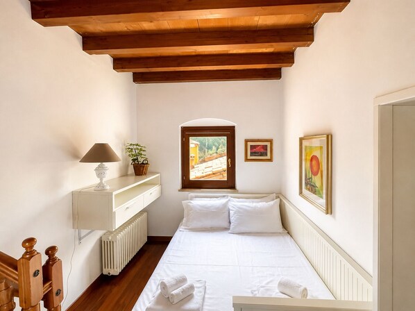 A narrow room with a bed, a radiator, a lamp, a plant, and two framed pictures.
