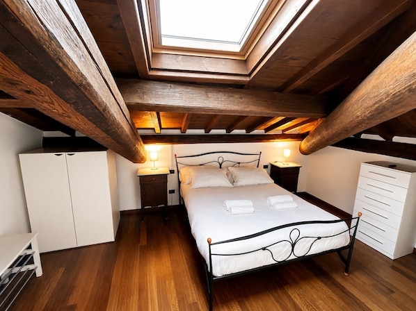 A bedroom with a skylight, wooden beams, a bed, bedside tables, a wardrobe, and a dresser.