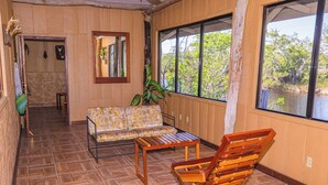 Um quarto com piso de madeira, um sofá, uma cadeira de madeira, uma mesinha, um espelho e uma janela com vista para árvores e água.