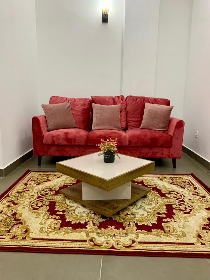 Un canapé rouge agrémenté de plusieurs coussins, une table basse blanche sur laquelle est posé un vase, et un tapis aux motifs rouges et dorés.