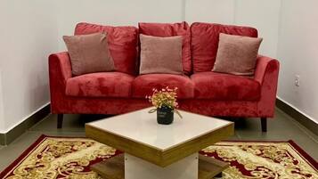 Un canapé rouge agrémenté de plusieurs coussins, une table basse blanche sur laquelle est posé un vase, et un tapis aux motifs rouges et dorés.