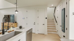 Una cocina moderna con lavavajillas, un lavamanos y una lámpara colgante. Se ve una escalera que sube a otro nivel.