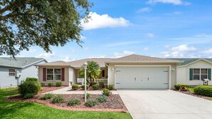 A single-story house with a front garage, a well-maintained lawn, and a small garden area with various plants and a palm tree. - Discover Your Perfect Summerhill Retreat with 2-Bed, 2-Bath, and Golf Cart! (The Villages)