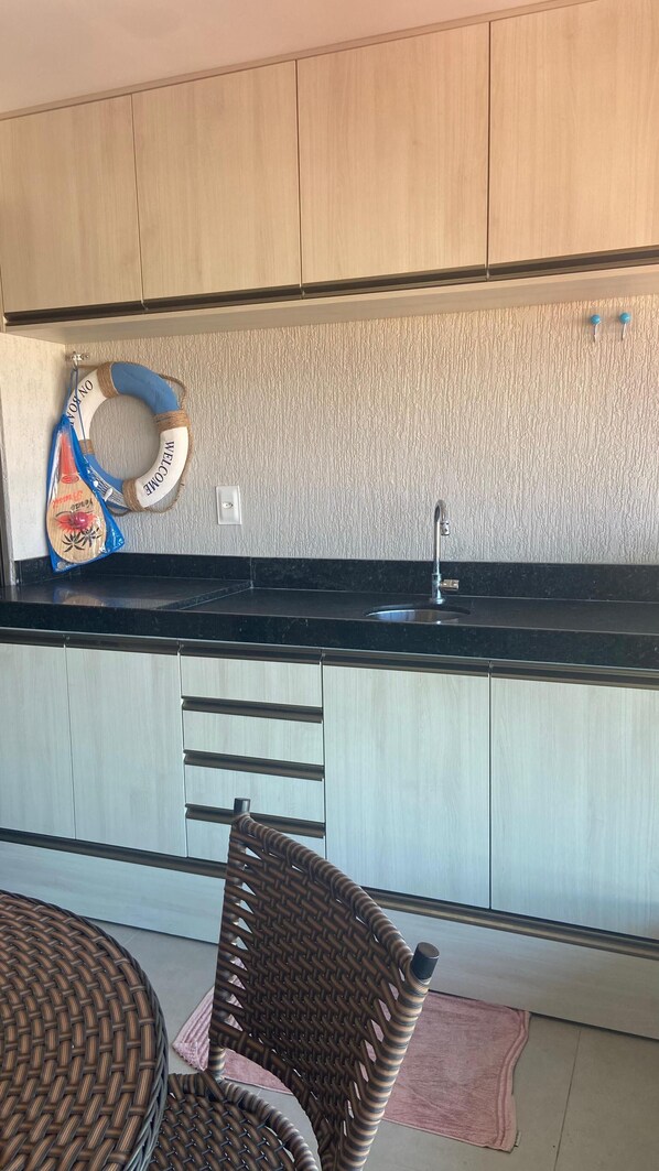 A kitchen with wooden cabinets, a black countertop, a sink, and a wicker chair. - Flat Principe Linda Vista Mar by LaPlaya (Ipojuca)