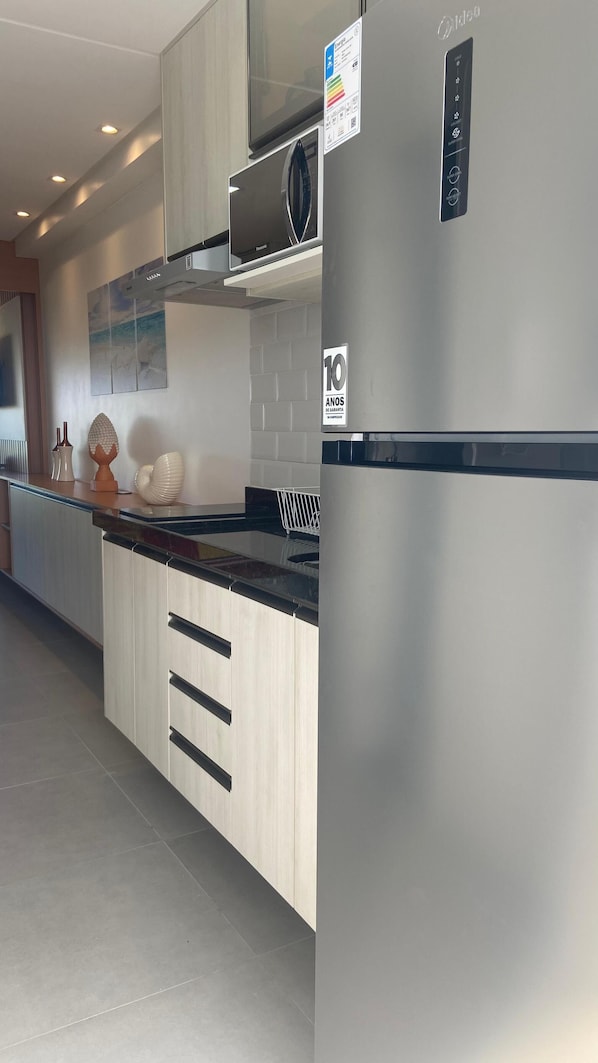A modern kitchen with stainless steel appliances, a microwave, and a refrigerator. There is a sign indicating a 10-year warranty on the refrigerator. The kitchen has a light wood finish and a black countertop. - Flat Principe Linda Vista Mar by LaPlaya (Ipojuca)