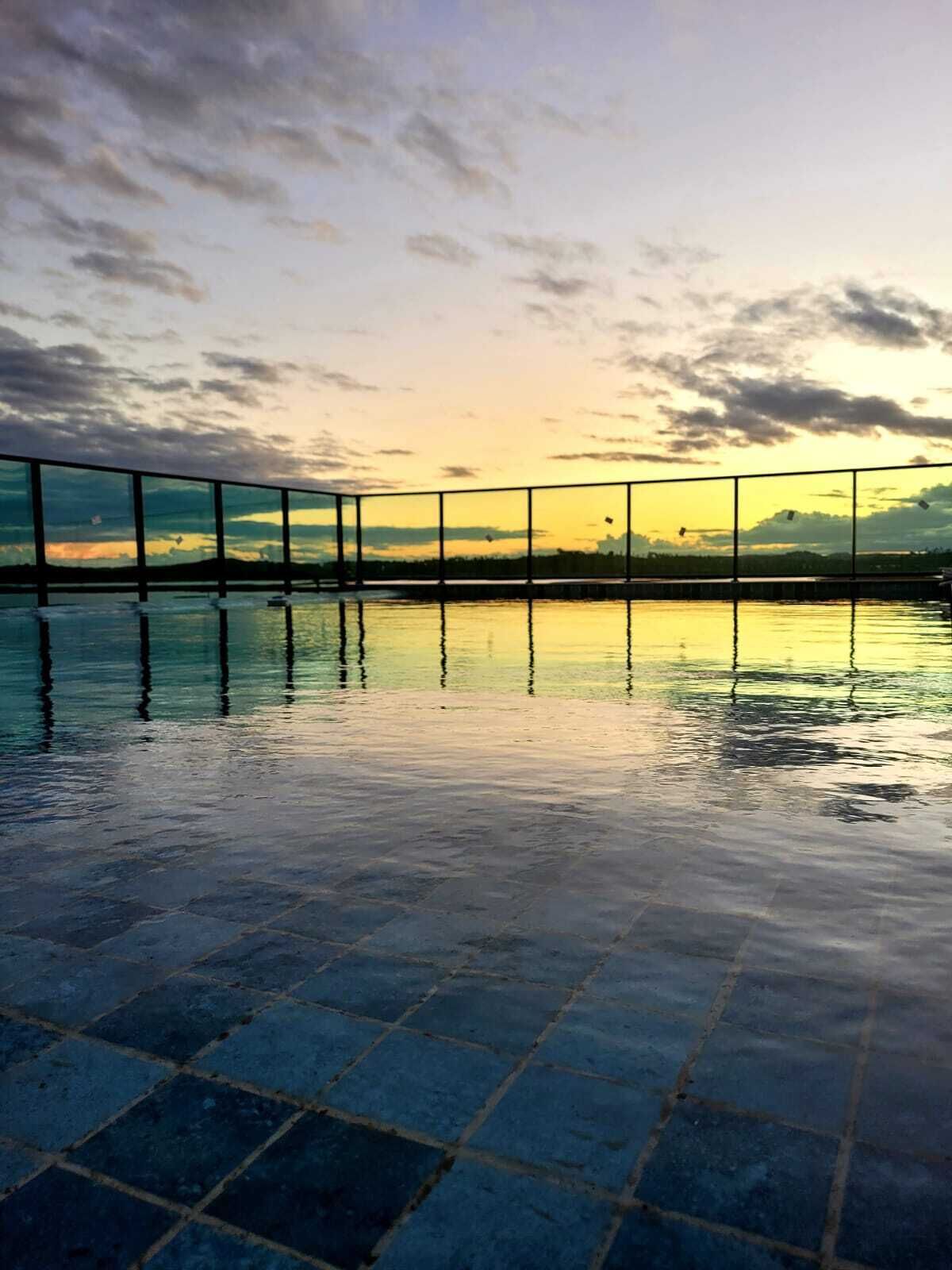 A tranquil outdoor pool area with a tiled floor and a railing overlooking a body of water, reflecting the sky.