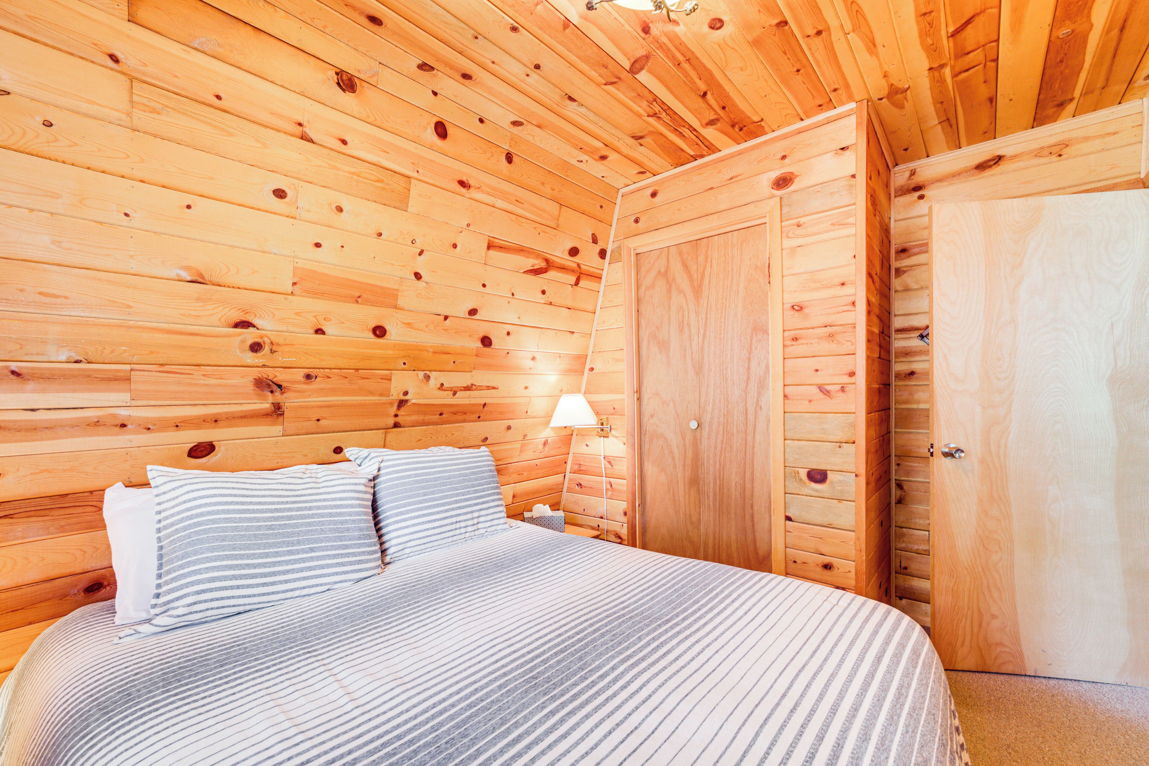 Una habitación de cabaña de madera con una cama, una mesita de noche, una lámpara y un armario.