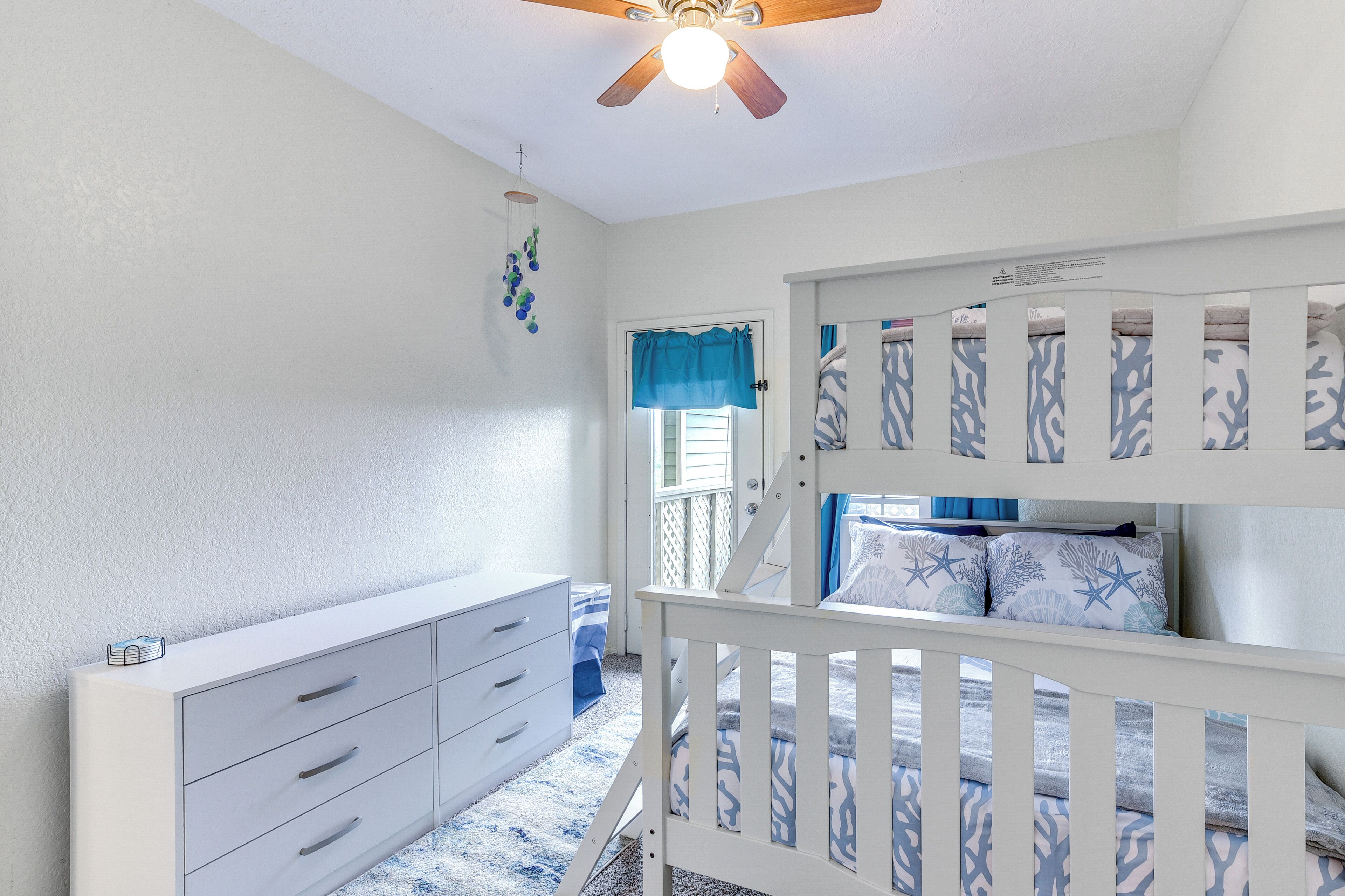 A child's bedroom with a bunk bed, a white dresser, a ceiling fan, and a window with blue curtains.