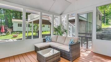 Een overdekt terras met een rietenbank, een glazen salontafel en uitzicht op een groen huis met een trap.