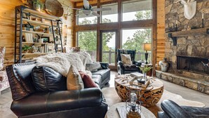 L’intérieur chaleureux d’un chalet en bois, avec une cheminée, des canapés en cuir, une bibliothèque et une grande fenêtre donnant sur le paysage extérieur.
