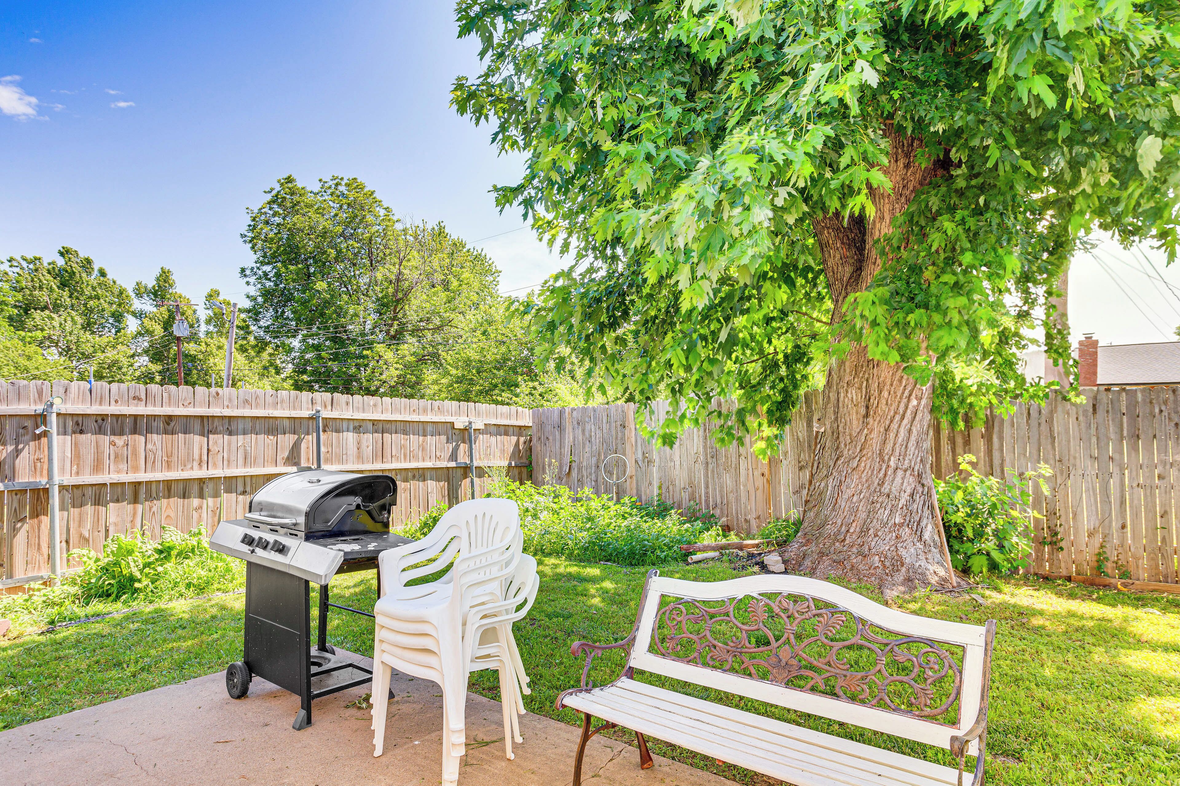 Ein Garten mit Grill, weißen Stühlen, einer Bank und einem großen Baum.