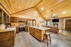 A spacious kitchen with wooden cabinets, a large island, and a stone fireplace. - Broken Bow Cabin w/ Private Hot Tub: 14 Mi to Lake (Broken Bow)