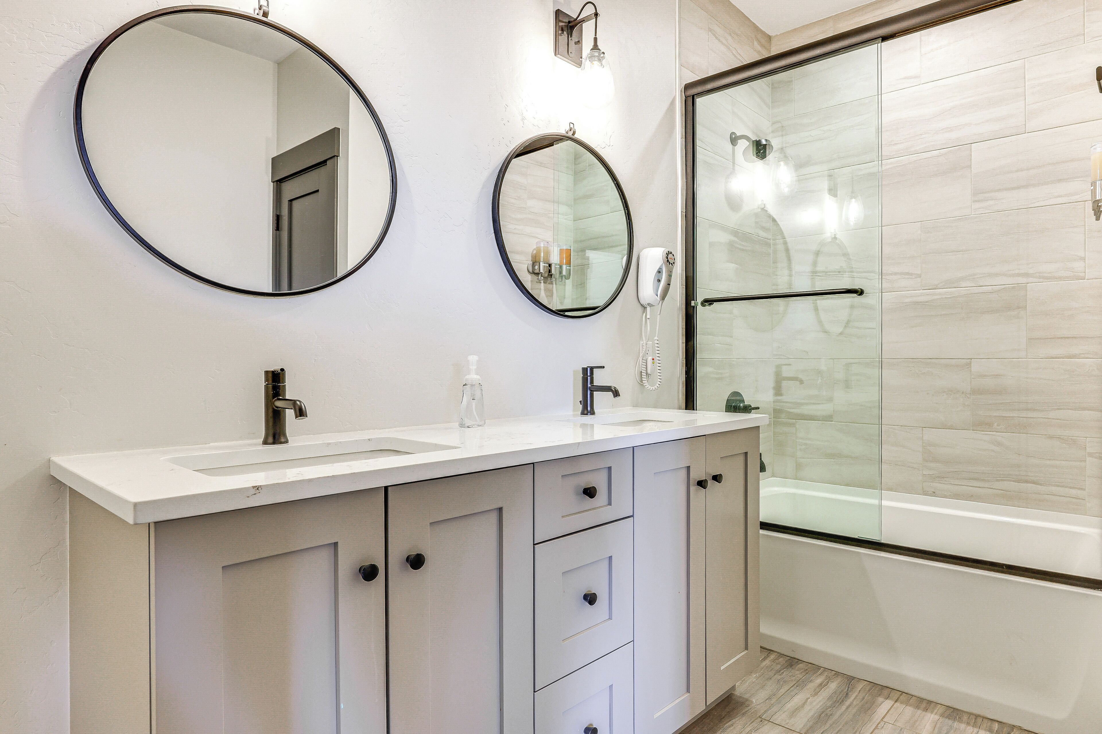 Un baño moderno con tocador blanco, dos espejos redondos, cabina de ducha de cristal y bañera.