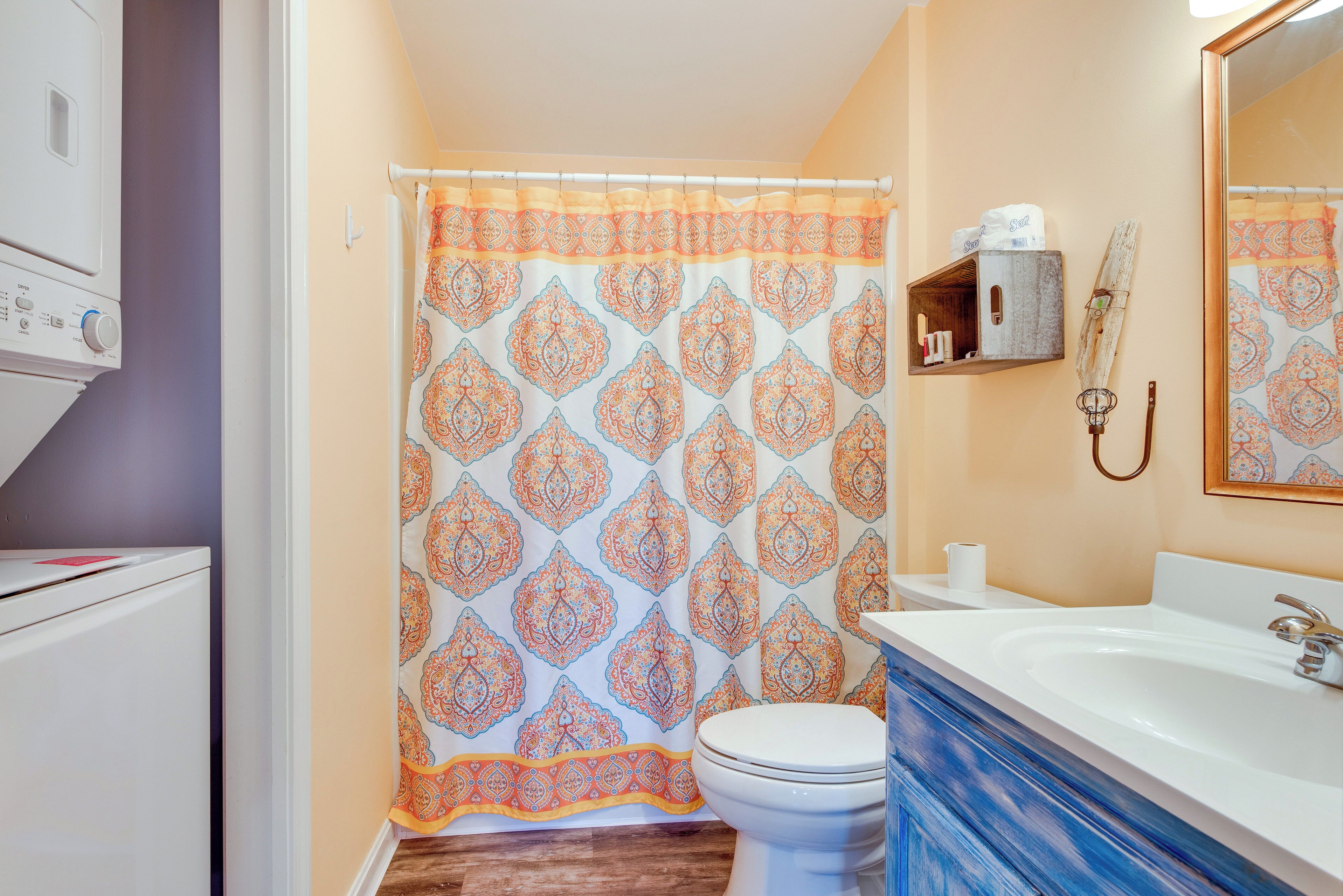 Un baño con cortina de ducha estampada, un inodoro blanco, un lavabo con espejo y un estante de madera con artículos de aseo.