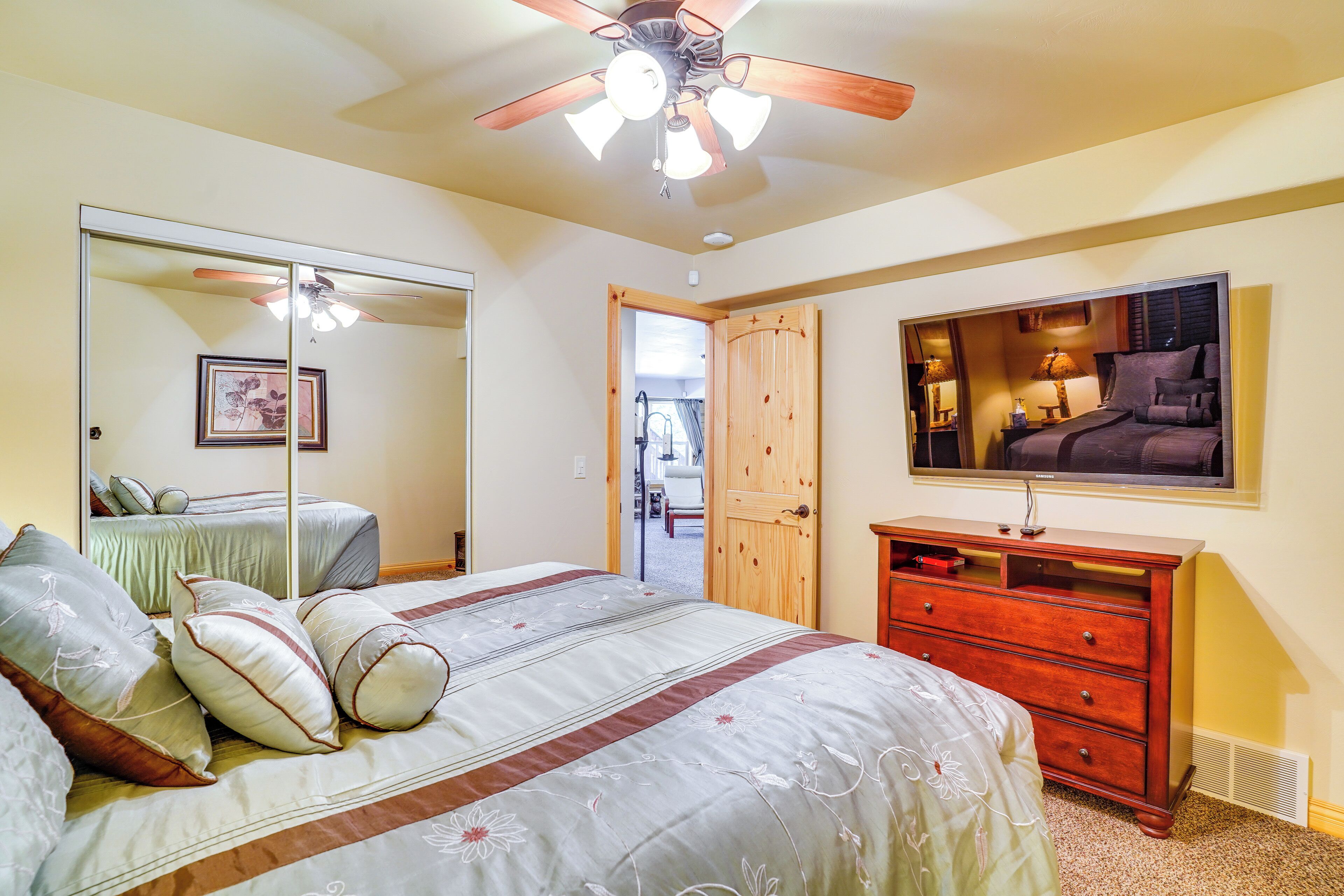 A bedroom with a bed, dresser, mirror, ceiling fan, and a door leading to another room.