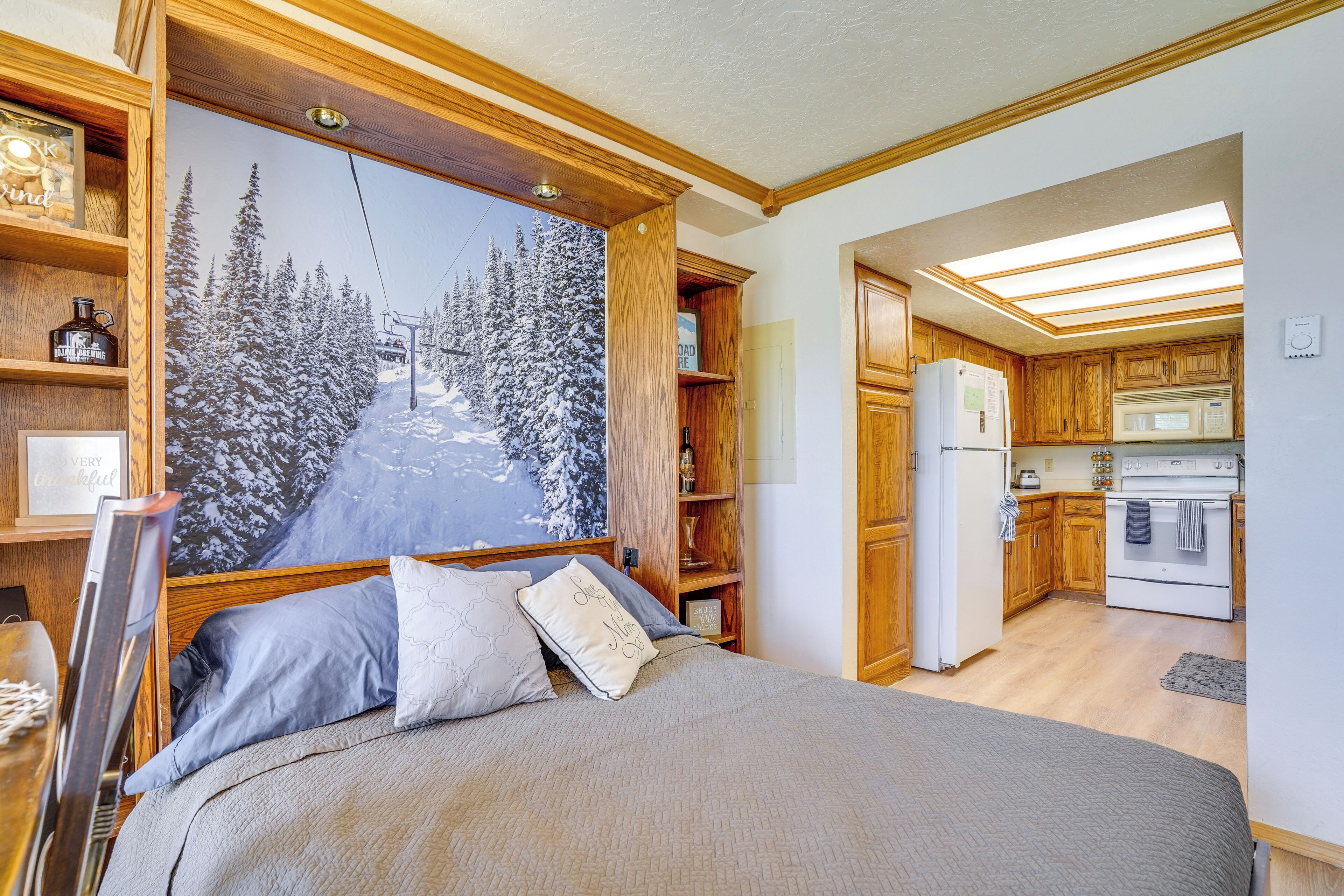 A bedroom with a large bed, wooden headboard, and a ski slope mural. Adjacent is a kitchen with wooden cabinets and a refrigerator.