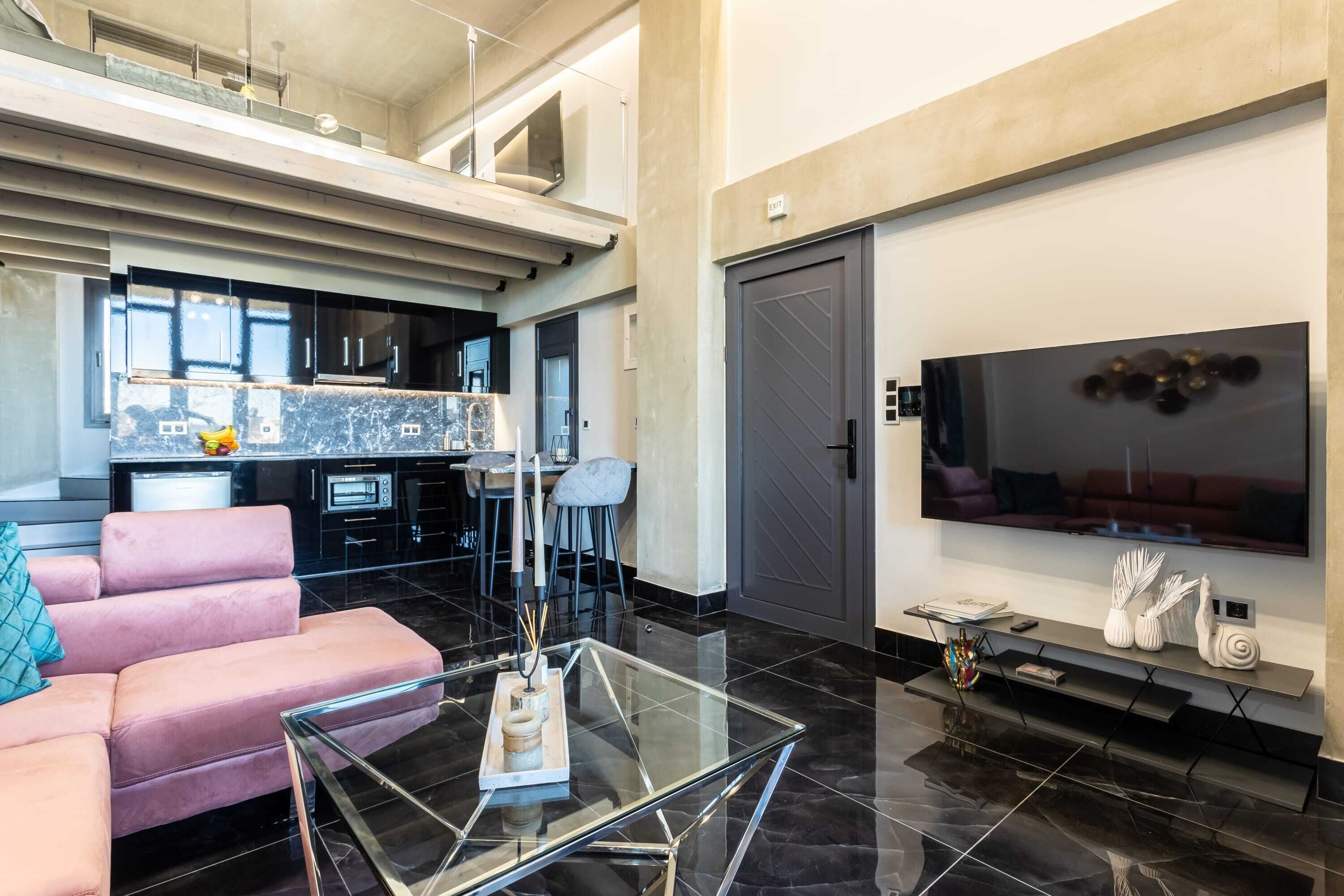 A modern living space with a glass coffee table, a pink sofa, a kitchen area, and a large flat-screen TV.