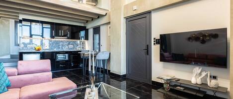 A modern living space with a glass coffee table, a pink sofa, a kitchen area, and a large flat-screen TV.