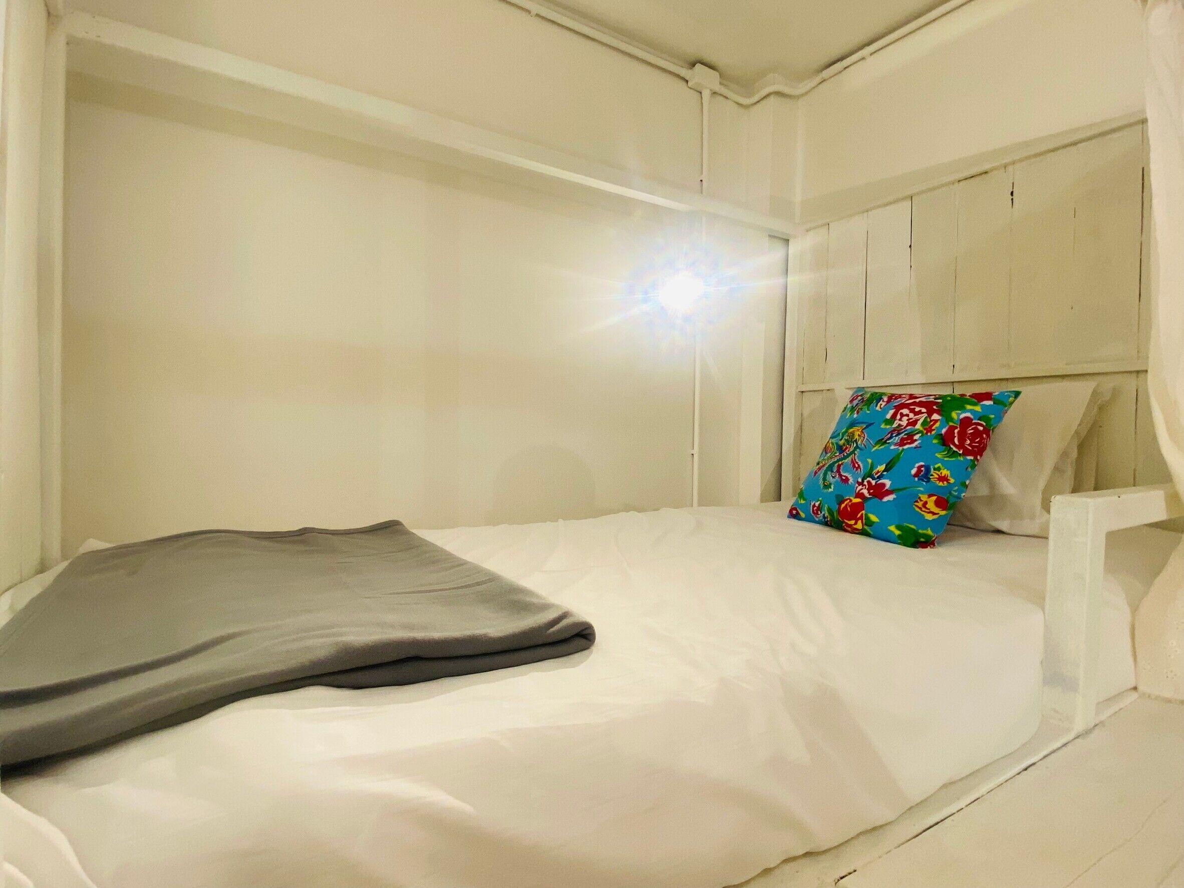 A single bed with white bedding and a colorful pillow, a grey blanket, and a white headboard.